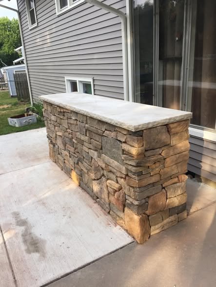 Outdoor stone bar and patio half-wall faced in stacked stone veneer with a chiseled-edge cap, set on a concrete patio slab